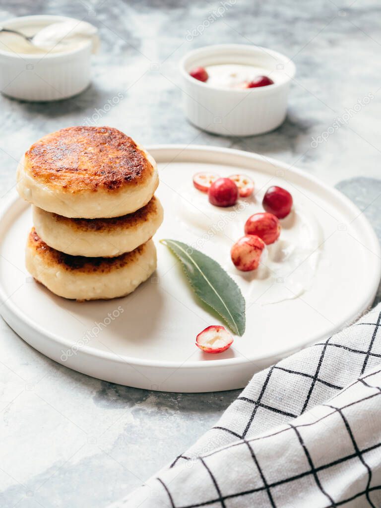 Cottage cheese pancakes on a table with sour cream and berries. Delicious and healthy cheesecakes with cranberry for breakfast. Yummy dessert. Vertical view, soft focus.