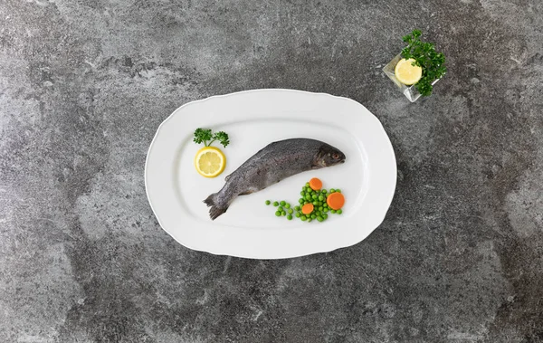 Limonlar, maydanozlar, bezelyeler ve havuçlarla dolu gri mermer bir tabakta tek bir gökkuşağı alabalığı.