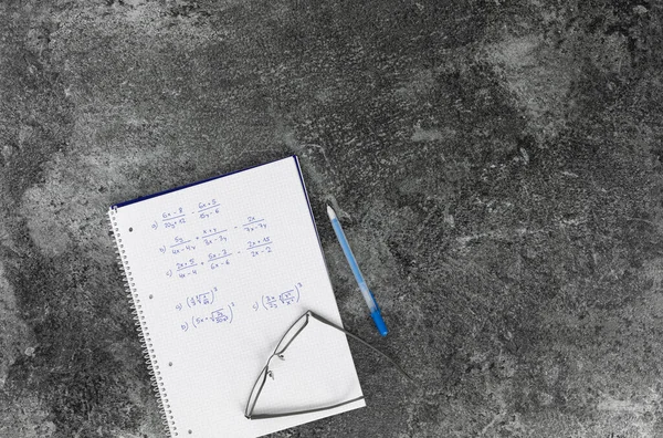 Matematik kitabı, aritmetik problemleri, gözlükler, mavi kalem ve gri mermer bir masa.