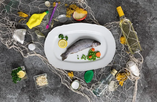 Gökkuşağı alabalıklı beyaz balık tabağı, bezelye, havuç, limon ve gri mermer bir masanın üzerindeki balık ağındaki çeşitli atıklar.