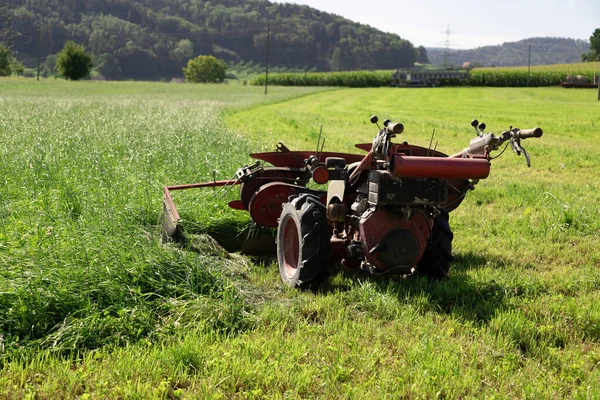 Çiftçinin çim biçmek için kullandığı büyük kırmızı iki el çim biçme makinesi, tarlada insan olmadan duruyor, yarısı biçilmiş, diğer tarafı ise mavi gökyüzünün önünde kesilmemiş yeşil çimlerle kaplı.