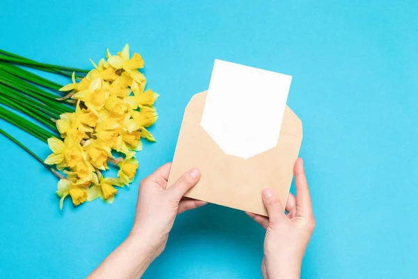Office desk, spring concept. Women\'s hands hold craft envelope with a postcard, fresh yellow daffodils on the background