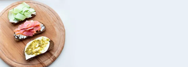 Morning breakfast of assorted sandwiches with different fillings on a wooden board. Fish sandwich with cheese and cucumber on the background. — Stock Photo, Image