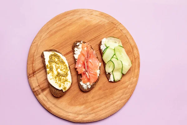 Petit déjeuner matinal de sandwichs assortis avec différentes garnitures sur une planche en bois. Sandwich au poisson avec fromage et concombre sur le fond . — Photo