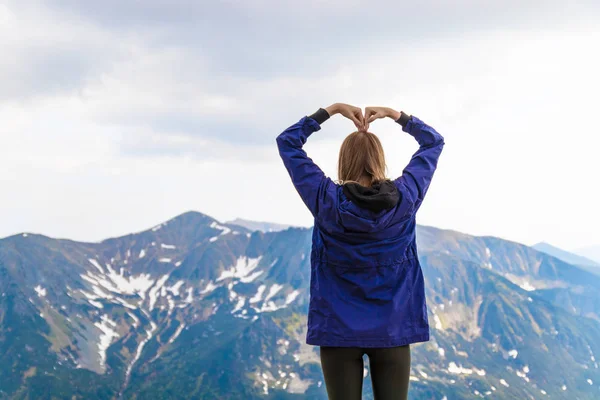 Młoda kobieta kształtuje serce z rąk, ciesząc się widokiem na piękne góry krajobrazu — Zdjęcie stockowe