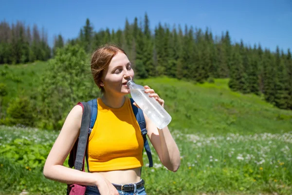 Female tourist girl in a yellow T-shirt and blue jeans shorts drink water from a plastic bottle on a background of green forest — Stock Photo, Image