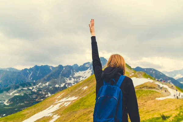 Blondynka podróżnik z niebieskim plecakiem podniósł rękę i ciesząc się zieloną górską scenerią, widok z tyłu — Zdjęcie stockowe