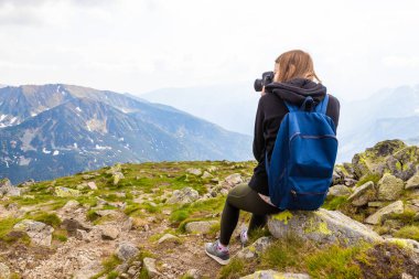 Genç bir sarışın mavi bir sırt çantası ile seyahat, bir dağın üstüne oturur ve yeşil dağ sahne resimleri alır. Geri görünüm