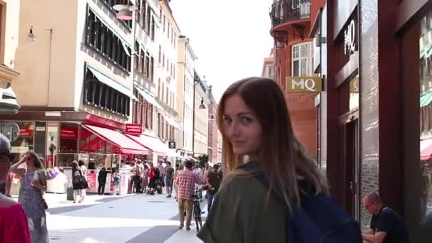 ストックホルム、スウェーデン - 2019年6月7日:明るいドレスを着たブロンドの女の子は、晴れた日に店や人々の間で通りを歩きます — ストック動画