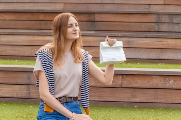 Молода красива жінка-працівник тримає паперовий обідній пакет, сидячи на дерев'яній лавці в парку. Обідня перерва — стокове фото