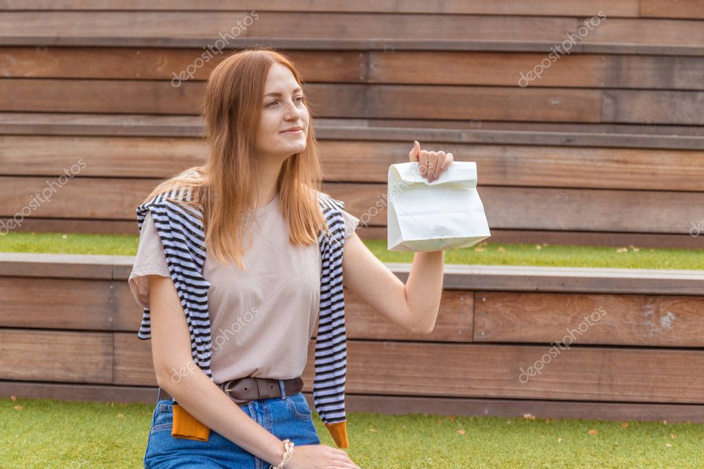 Smiling blonde student girl in t-shirt and blue jeans is sitting on a wooden bench holding a doggy bag on a sunny day in the park. Lunch break. Empty space for design