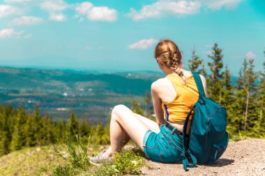 Sırt çantalı ve gözlüklü sarışın kız bir dağda oturur ve güneşli bir günde dağların güzel tepelerinin tadını çıkarır..
