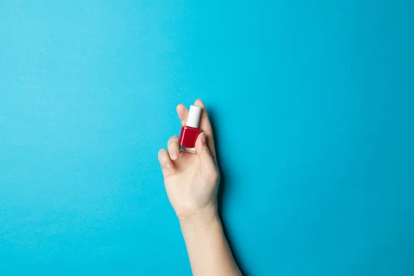 Mão de mulheres mantém uma manicura de vidro uma garrafa com verniz vermelho em um fundo azul escuro claro . — Fotografia de Stock
