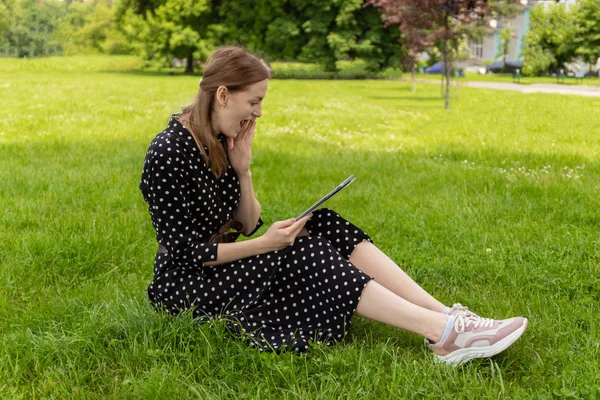 远程教育。穿着夏装的年轻金发学生坐在草地上，用平板电脑. — 图库照片