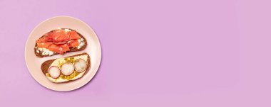 Mor bir arka plan üzerinde pembe seramik plaka üzerinde kırmızı balık, beyaz peynir salatalık ve pesto ile taze sandviç