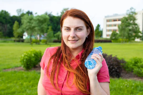 Young girl in bright summer clothes holds a plastic bottle of water on the background of the park