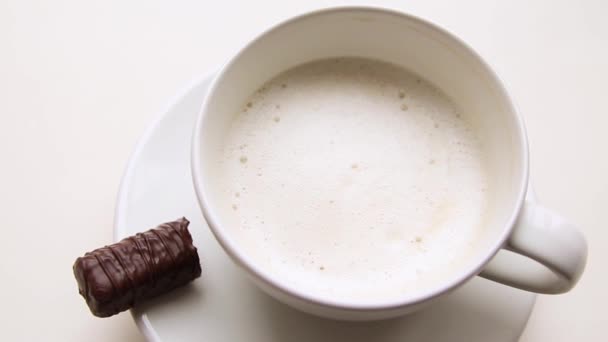 Свежеприготовленный капучино в белой чашке с шоколадной конфеткой на белом столе — стоковое видео