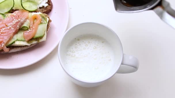 Barista el çırpılmış süt ile seramik bir fincan içine kahve döker. Arka planda salatalık ve balık ile bir sandviç. — Stok video
