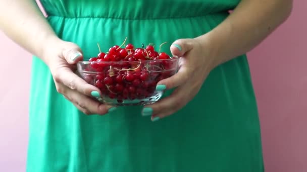 A girl in a green dress holds in her hands a glass bowl with red ripe currants on a pink background. Summer berry concept. — Stock Video