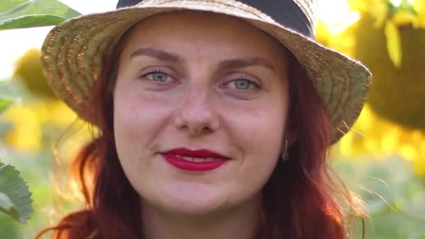 Рудоволоса дівчина в солом'яному капелюсі і червона помада посміхається, дивлячись на камеру. Величезне жовте поле соняшників — стокове відео