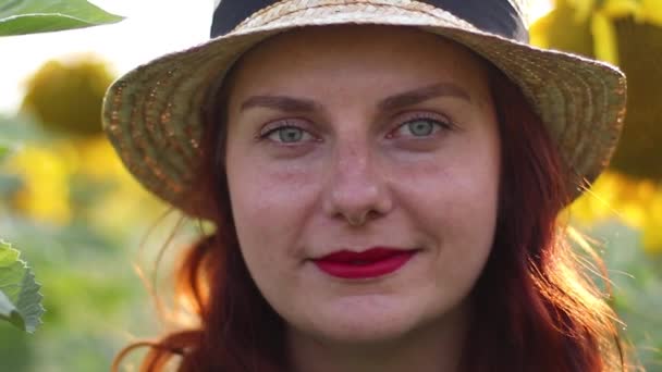 Chica pelirroja con sombrero de paja y lápiz labial rojo sonríe mientras mira a la cámara. Campo enorme de girasoles amarillos — Vídeos de Stock