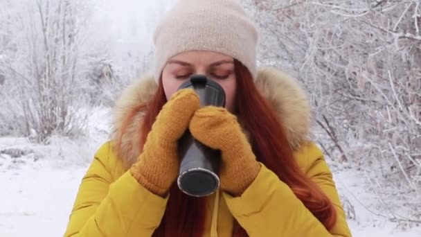 Jonge roodharige meisje in een warme gebreide muts en een gele winterjas drinkt een warm drankje uit een metalic mok tegen de achtergrond van een winter landschap. — Stockvideo