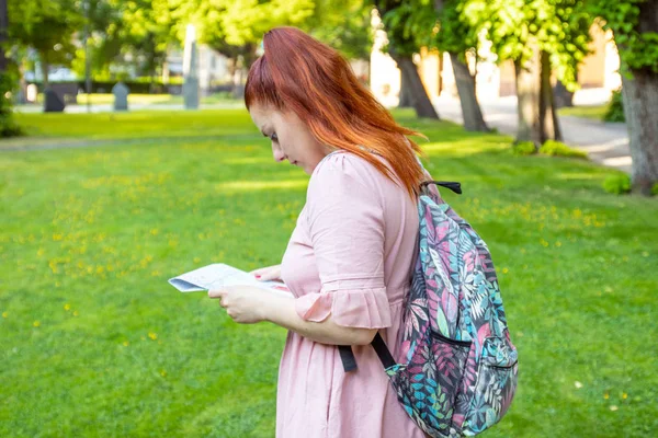 一个红头发的女孩，穿着粉红色的裙子，拿着一张地图，在公园的绿草上探索城市周围的旅行路线。. — 图库照片