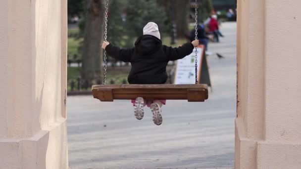 Baksidan på en liten flicka i vit hatt och svart rock rullar ensam på swing i en lekplats. Långsam moské. — Stockvideo