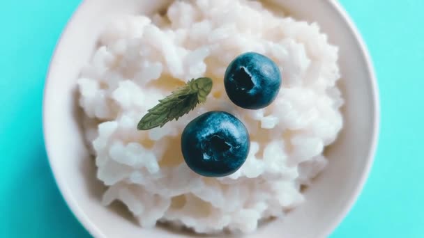 Плоская каша из рисового молока с черникой и мятой на голубом фоне. Вкусный здоровый завтрак — стоковое видео