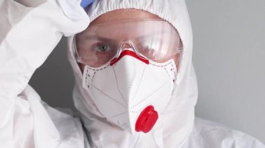 Tıbbi koruyucu maskeli, gözlüklü ve tulumlu yorgun bir doktorun yakın portresi.