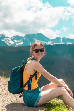 Sırt çantalı ve gözlüklü sarışın kız bir dağda oturur ve güneşli bir günde dağların güzel tepelerinin tadını çıkarır..