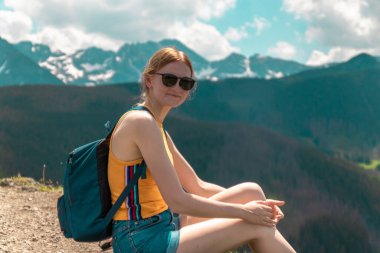 Sırt çantalı ve gözlüklü sarışın kız bir dağda oturur ve güneşli bir günde dağların güzel tepelerinin tadını çıkarır..