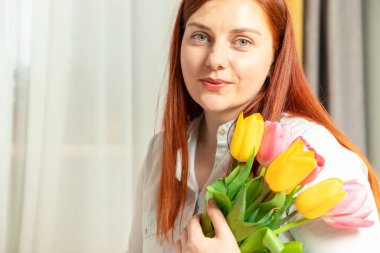 Mavi pamuklu bluzlu güzel kızıl saçlı kız ellerinde taze lale buketiyle gri bir arka planda.