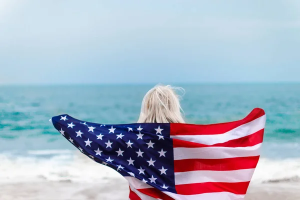 Biała blondynka trzymająca amerykańską flagę opierającą się o plecy i patrzącą na morze — Zdjęcie stockowe