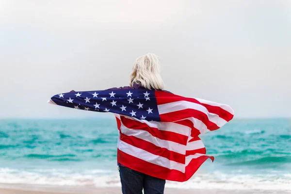 Biała blondynka stojąca całkowicie owinięta w amerykańską flagę i patrząca na morze. Flaga USA na Dzień Niepodległości — Zdjęcie stockowe