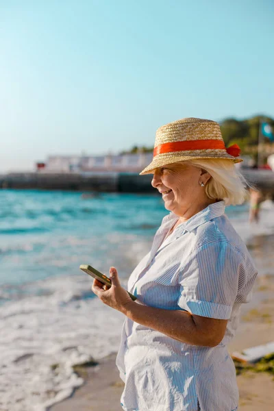 Summer holiday background. Caucasian blond woman in straw hat and striped shirt makes photo by smartphone of the sea on a sunny day.