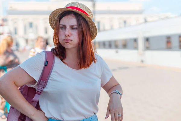 Una joven cansada en un sombrero con una mochila y una maleta se para en la estación esperando el transporte público — Foto de Stock
