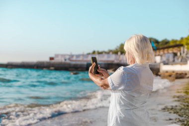 Mutlu bir kadın tatilde, deniz sahilinde kamerayla fotoğraf çekiyor. Akıllı telefonuyla, boş zamanların ve yaz tatilinin tadını çıkarıyor.