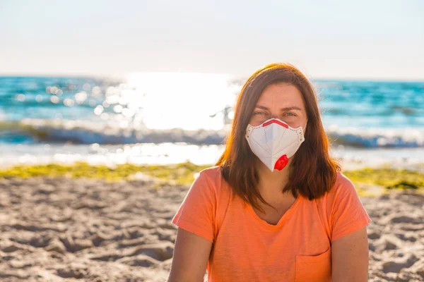 Fiatal nő védő maszkban ül a tengerparton a tenger mellett. Új szabályok az arcfedő maszk viselésére nyilvános helyeken a koronavírus miatt COVID 19. — Stock Fotó