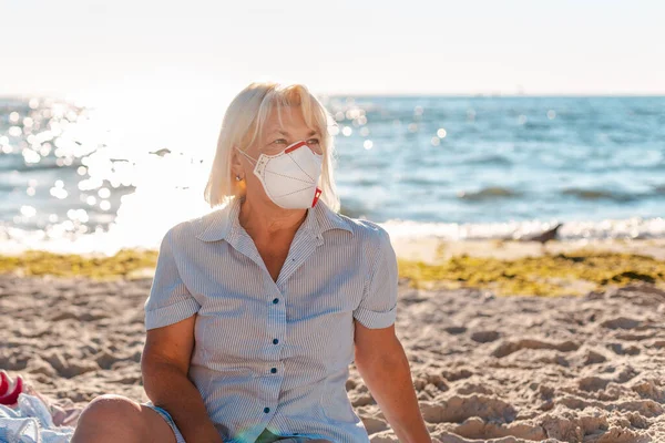 Szőke, védő maszkban és ingben, a kamerába néz, a parton ül, közel a strandhoz. — Stock Fotó