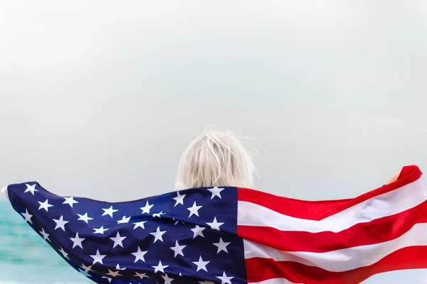 Biała blondynka stojąca całkowicie owinięta w amerykańską flagę i patrząca na morze. Flaga USA na Dzień Niepodległości — Zdjęcie stockowe
