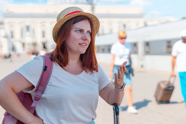 Tren istasyonunda sırt çantası ve bavul taşıyan şapkalı yorgun, kızıl saçlı bir kız. — Stok fotoğraf
