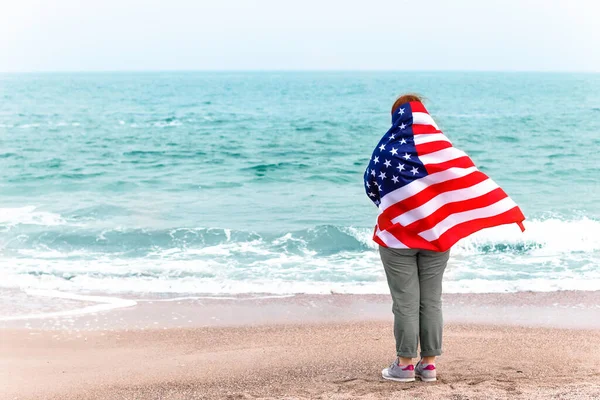 Widok z tyłu młodej dziewczyny z amerykańską flagą patrzy na morze — Zdjęcie stockowe