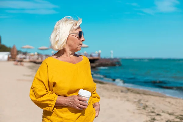 Blond kvinna som håller papper kopp kaffe med henne på havet — Stockfoto