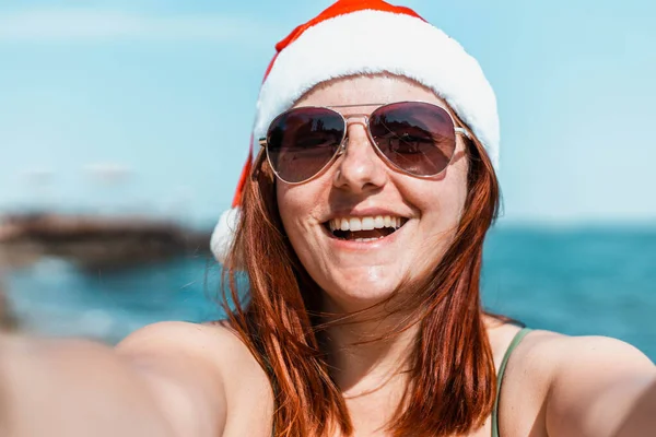 Piękna szczęśliwa dziewczyna w santa kapelusz i strój kąpielowy sprawia selfie i korzystających zimowe wakacje wypad na tropikalnej plaży. — Zdjęcie stockowe