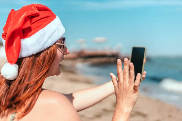 Jong mooi meisje in een rood kerstman hoed en badpak maakt een selfie op de kust — Stockfoto
