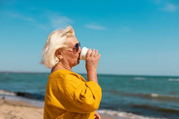Adult blond woman in sunglasses and yellow blouse drinks coffee from a paper cup on the seashore on a sunny day. Coffee to go — Stock Photo, Image