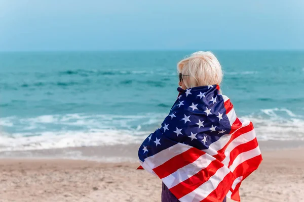 Biała blondynka zawróciła trzymając amerykańską flagę i zostaje na plaży w pobliżu morza. — Zdjęcie stockowe