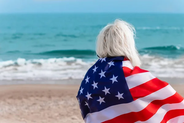 Biała blondynka trzymająca amerykańską flagę opierającą się o plecy i patrzącą na morze — Zdjęcie stockowe