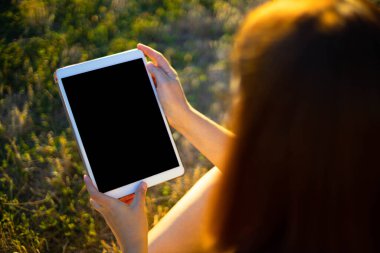 Model laptop. Parktaki yeşil çimlerde boş ekranlı tablet tutan genç bir kadın.